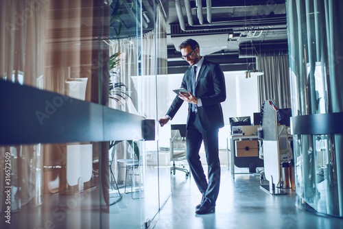 Fotografía Full length of attractive Caucasian bearded businessman in suit and with eyeglasses holding tablet and entering his office