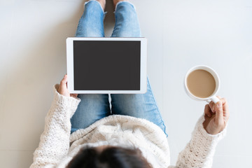 Smiling pretty woman in white sweater using tablet computer while holding a cup of coffee sitting in the living room at home. Top view, copy space.