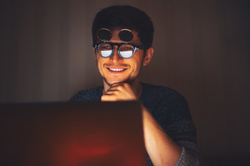 Canvas Print - Night portrait of young happy man wearing round sunglasses, looking in laptop in dark room at home.