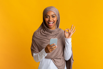 Wall Mural - Black muslim girl in hijab using smartphone and showing ok gesture