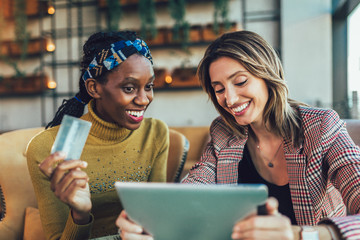 Two woman using tablet and credit card together inside cafe.