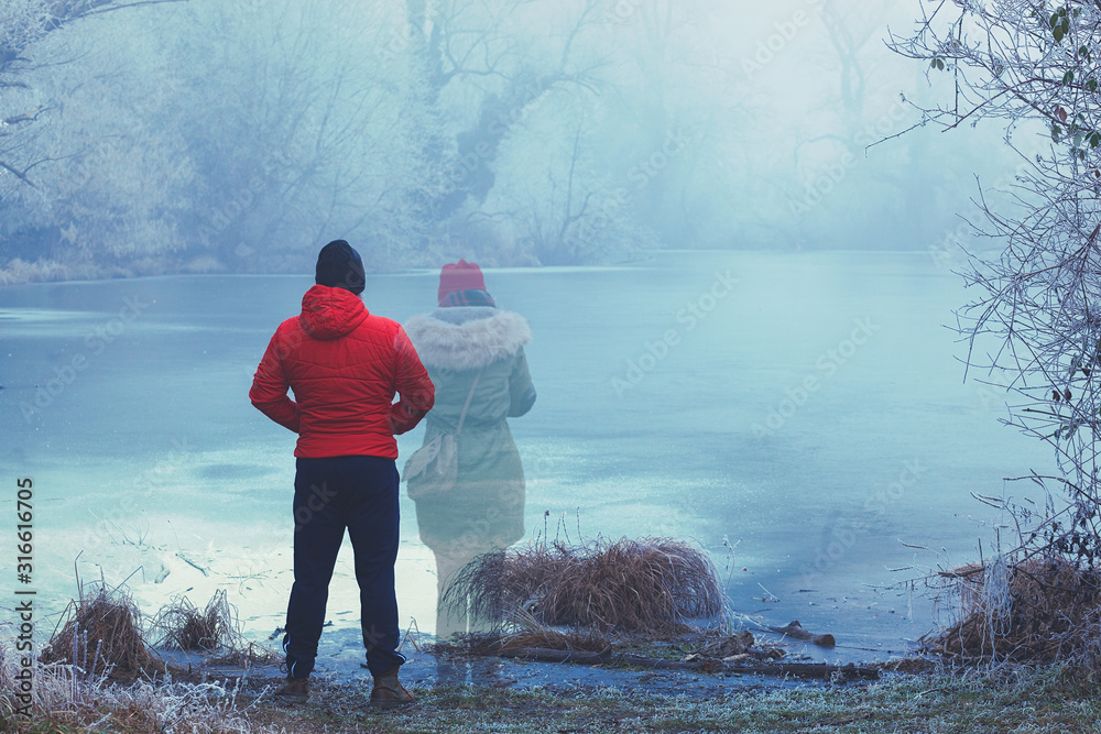 Lonely man in red jacket standing by the lake in winter, with transparent woman figure standing next to him - obrazy, fototapety, plakaty 