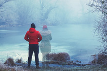 Lonely Man In Red Jacket Standing By The Lake In Winter, With Transparent Woman Figure Standing Next To Him