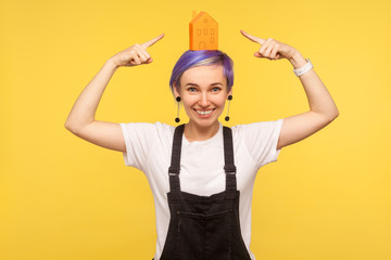 Portrait of happy modern fashionable hipster girl with violet short hair in denim overalls pointing at orange paper house on her head and smiling at camera. studio shot isolated on yellow background