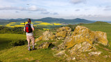 Fototapeta Na ścianę - A hiker with a backpack near the city of Kassel on a mountain. The stones have the name Helfensteine. Below is an airfield for gliders. Rape fields bloom in the distance.