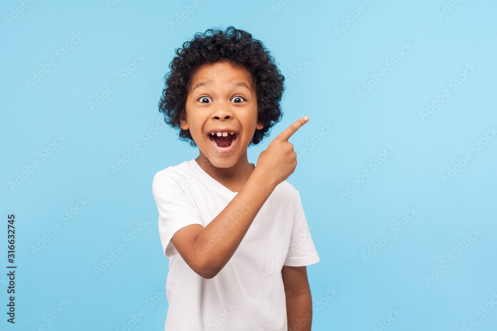 Wow look, advertise here! Portrait of amazed cute little boy with curly hair pointing to empty place on background, surprised preschooler showing copy space for promotional ad. indoor studio shot - obrazy, fototapety, plakaty 