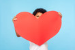 © khosrork - Cute shy adorable little boy peeking out from big red paper heart, unrecognizable child holding symbol of love, charity, looking with interest at camera. indoor studio shot isolated on blue background