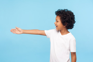 Profile of polite cheerful preschool boy with curly hair in white T-shirt giving hand to handshake, gullible trusting child meeting with friendly smile. indoor studio shot isolated on blue background