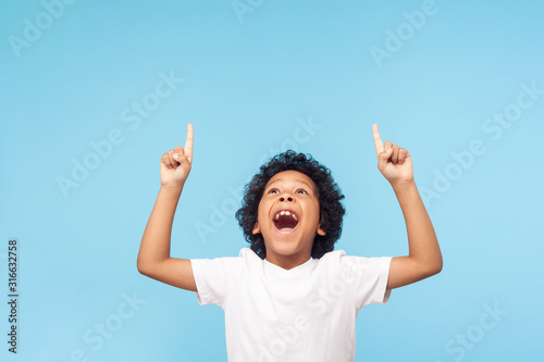 Cuadros en Lienzo Wow, look up! Portrait of amazed little boy pointing up to empty place on blue background, expressing shock surprise with wide open mouth and showing copy space for advertisement