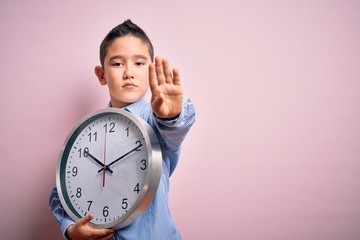 Poster - Young little boy kid holding big minute clock over isolated pink background with open hand doing stop sign with serious and confident expression, defense gesture