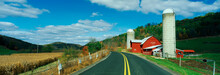 This Is A Country Road With A Farm On The Right Hand Side Of The Road. It Has A Red Barn And Silo.