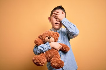 Canvas Print - Young little boy kid hugging teddy bear stuffed animal over yellow background stressed with hand on head, shocked with shame and surprise face, angry and frustrated. Fear and upset for mistake.