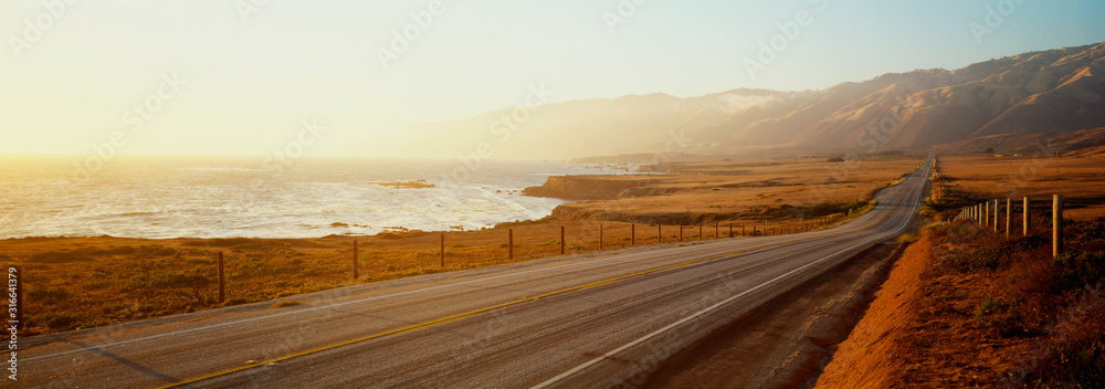 This is Route 1also known as the Pacific Coast Highway. The road is situated next to the ocean with the mountains in the distance. The road goes off into infinity into the sunset. - obrazy, fototapety, plakaty 