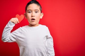 Canvas Print - Young little boy kid holding heart paper shape over isolated red background scared in shock with a surprise face, afraid and excited with fear expression