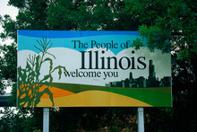 This Is A Road Sign That Says, The People Of Illinois Welcome You. There Is A Stalk Of Corn On One Side Of The Sign, And The City Skyline Of Chicago On The Other Side. It Is Set Near Green Trees.