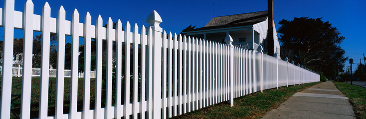 This is a close up of a white picket fence and a typical looking suburban house. We see the sidewalk that runs past it on the right hand side of the image.