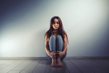 a young sad woman sits disheveled on the floor of a room. there is a light wall in the background.hu
