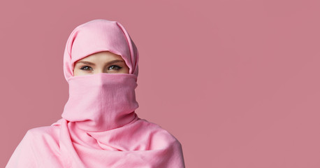 Portrait of young Muslim arabian woman wearing colorful hijab against pink background. Points finger to side. Space for text