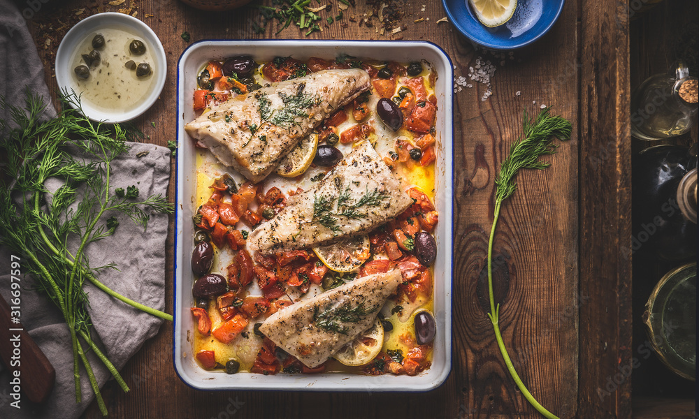 Tasty Bass fish fillets in Mediterranean sauce with tomatoes, olives and capers in baking pan on rustic wooden background with ingredients. Top view - obrazy, fototapety, plakaty 