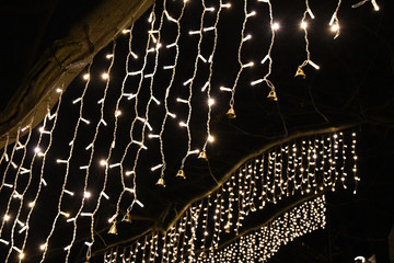 Wall Mural - Abstract background. Rows of Christmas illuminated decorations with shining garlands and small shiny metal bells on black night background. Glowing hanging lights. New Year celebration decor.