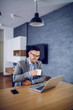 © dusanpetkovic1 - Smiling bearded caucasian businessman in suit and with eyeglasses sitting at dining table, drinking fresh morning coffee and looking at laptop. On table are smart phone and laptop.