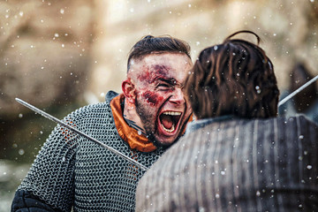 Two warriors in armor with weapons fighting with swords. Close up of emotions on the face of a warrior smeared with blood