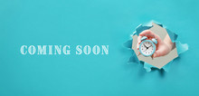 Coming Soon Text On Blue Background. Alarm Clock In Hand. Concept Of Announcement, Opening