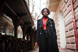 Fototapeta Krajobraz - Portrait of a curly haired african woman wearing fashionable black coat and red turtleneck walking outdoor.