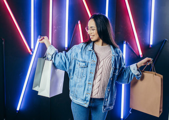 Young girl in blue denim jacket with shopping bags on neon lamps background.