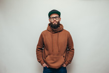 Handsome Hipster Guy With Beard Wearing Brown Blank Hoodie Or Hoody And Black Cap With Space For Your Logo Or Design On White Background. Mockup For Print