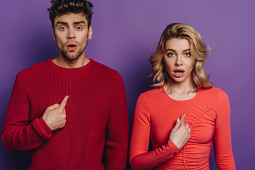 surprised man and woman pointing with fingers at themselves while looking at camera on purple background