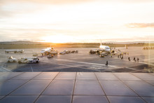 Airplanes On The Airport Runway. Operators Loading And Unloading And People Boarding The Planes. Airport At Sunset