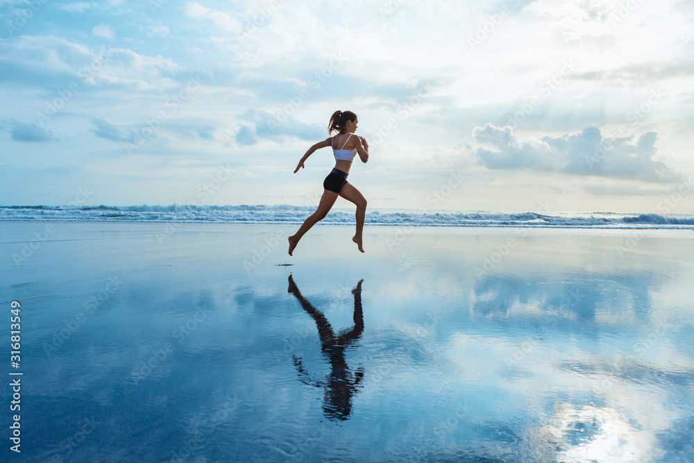 Barefoot young girl with slim body running along sea surf by water pool to keep fit and burning fat. Beach background with blue sky. Woman fitness, jogging sports activity on summer family vacation. - obrazy, fototapety, plakaty 