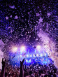 Menschen feiern vor einer Bühne auf einem Musikfestival mit Konfetti und bunten Scheinwerfern