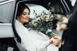 Bride with flower bouquet in car. Pretty woman wearing white wedding dress and tiara with veil. The bride gets out of the car and the groom extends a hand. Bride with wedding makeup and hairstyle
