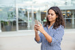 © Mangostar - Smiling woman using smartphone on street. Beautiful cheerful young woman standing outside modern building and using mobile phone. Technology concept