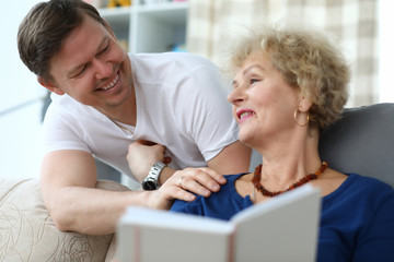 Portrait of happy cheerful relatives smiling at home. Smiling adult man having fun with aged mom. Cheerful woman enjoying reading book at home. Friendly family concept