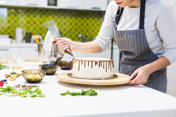 Wall Mural - A confectioner squeezes liquid chocolate from a pastry bag onto a white cream biscuit cake on a wooden stand. The concept of homemade pastry, cooking cakes.