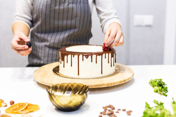 Wall Mural - Confectioner decorates with berries a biscuit cake with white cream and chocolate. Cake stands on a wooden stand on a white table. The concept of homemade pastry, cooking cakes.