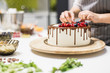 © xartproduction - Confectioner decorates with berries a biscuit cake with white cream and chocolate. Cake stands on a wooden stand on a white table. The concept of homemade pastry, cooking cakes.