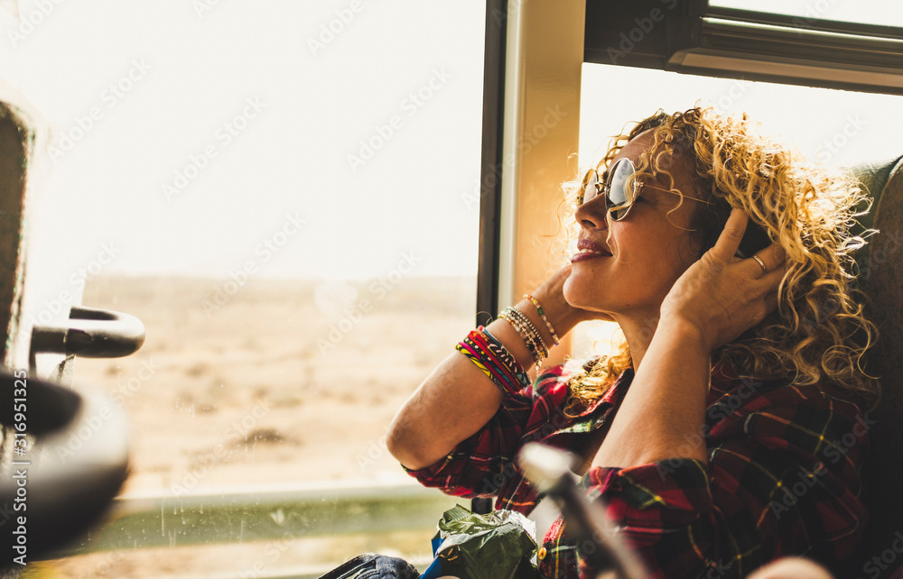 Beautiful blonde adult woman enjoying the travel on bus or train listening music with headphones - wanderlust lifestyle and happy people doing trip - happy people and transport - obrazy, fototapety, plakaty 
