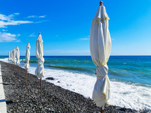 Eight White Beach Umbrella Stands In A Line Along The Shore Next To The Sea On Which The Wind Made Waves Amid A Bright Sky