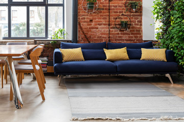 Interior of living room with comfortable navy sofa with yellow pillows and vertical garden. Red brick wall and big window in industrial loft apartment. Stylish lounge.