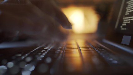 Fast motion close-up shot of laptop, hands and fingers of unrecognizable female programmer typing on keyboard late at night, illuminated by lamp in background, and lines of symbols running on screen