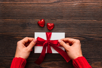 White present with a red ribbon in woman's hands and two chocolate sweets in heart shape on a dark wooden background. Valentine day concept. Copyspace.