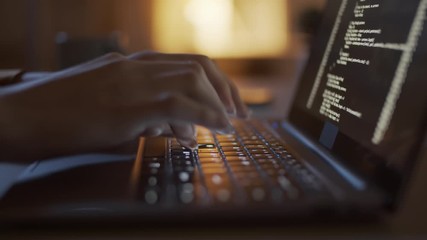 Close-up shot of hands of anonymous woman sitting at desk late at night, typing computer code on laptop keyboard, lines of digital symbols running on screen, and table lamp in background