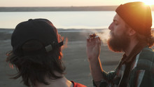 Two Friends Smoking Joint With Weed At Seashore At Sunset