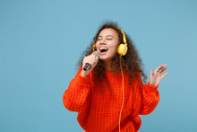 Funny Young African American Girl In Orange Knitted Clothes Isolated On Pastel Blue Background. People Lifestyle Concept. Mock Up Copy Space. Listen Music With Headphones, Sing Song In Microphone.