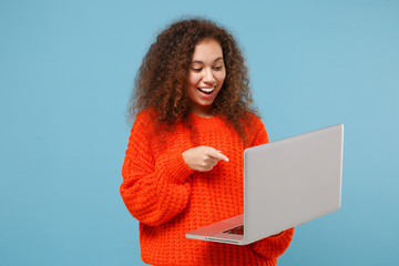 Funny young african american girl in casual orange knitted clothes isolated on pastel blue background in studio. People lifestyle concept. Mock up copy space. Point index finger on laptop pc computer.