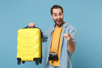 Excited traveler tourist man in yellow clothes with photo camera isolated on blue background. Male passenger traveling abroad on weekends. Air flight journey. Holding suitcase point finger on camera.